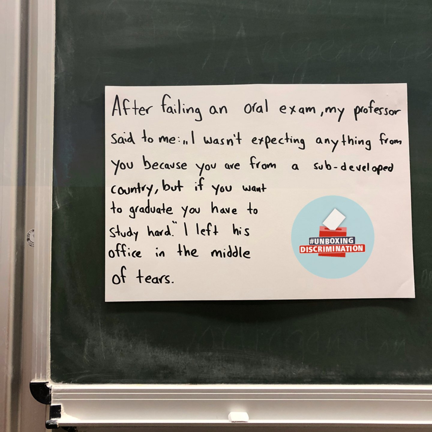 A white poster hangs on a chalkboard. Next to the logo of the campaign #unboxingdiscrimination is handwritten: "After failing an oral exam the professor said to me “ I was not expecting anything from you because you are from an sub-developed country, but if you want to graduate you have to study hard” I left his office in the middle of tears."