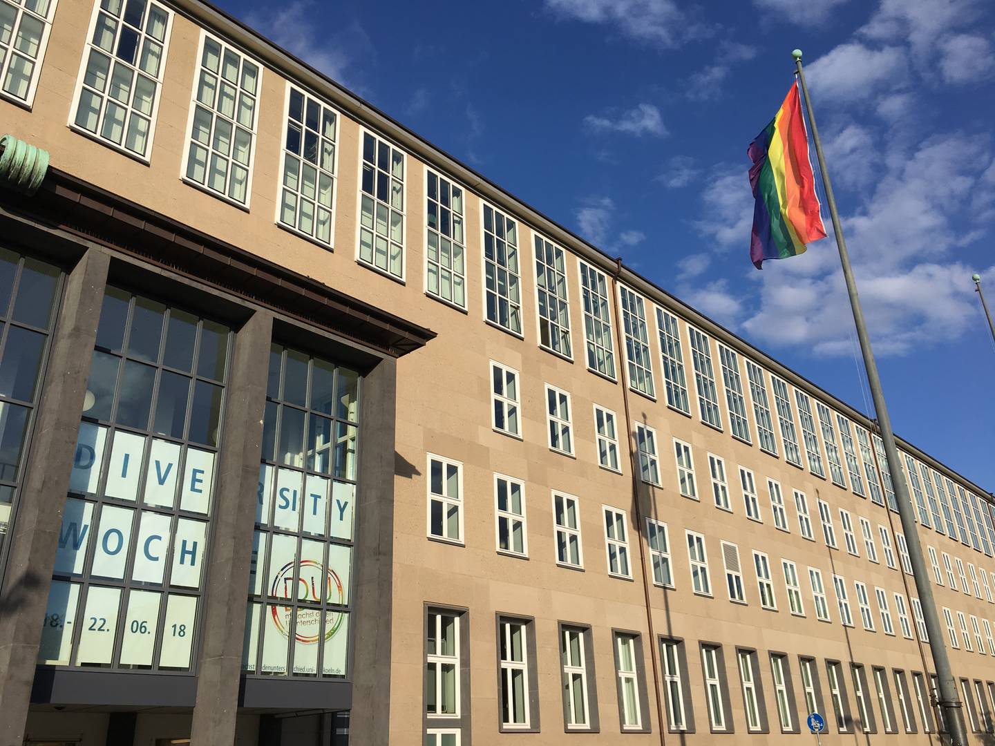 Auf dem Bild ist eine gehisste Regenbogenflagge zu sehen, welche vor blauem Himmel im Wind weht. Im Hintergrund sieht man das Hauptgebäude der Universität zu Köln.