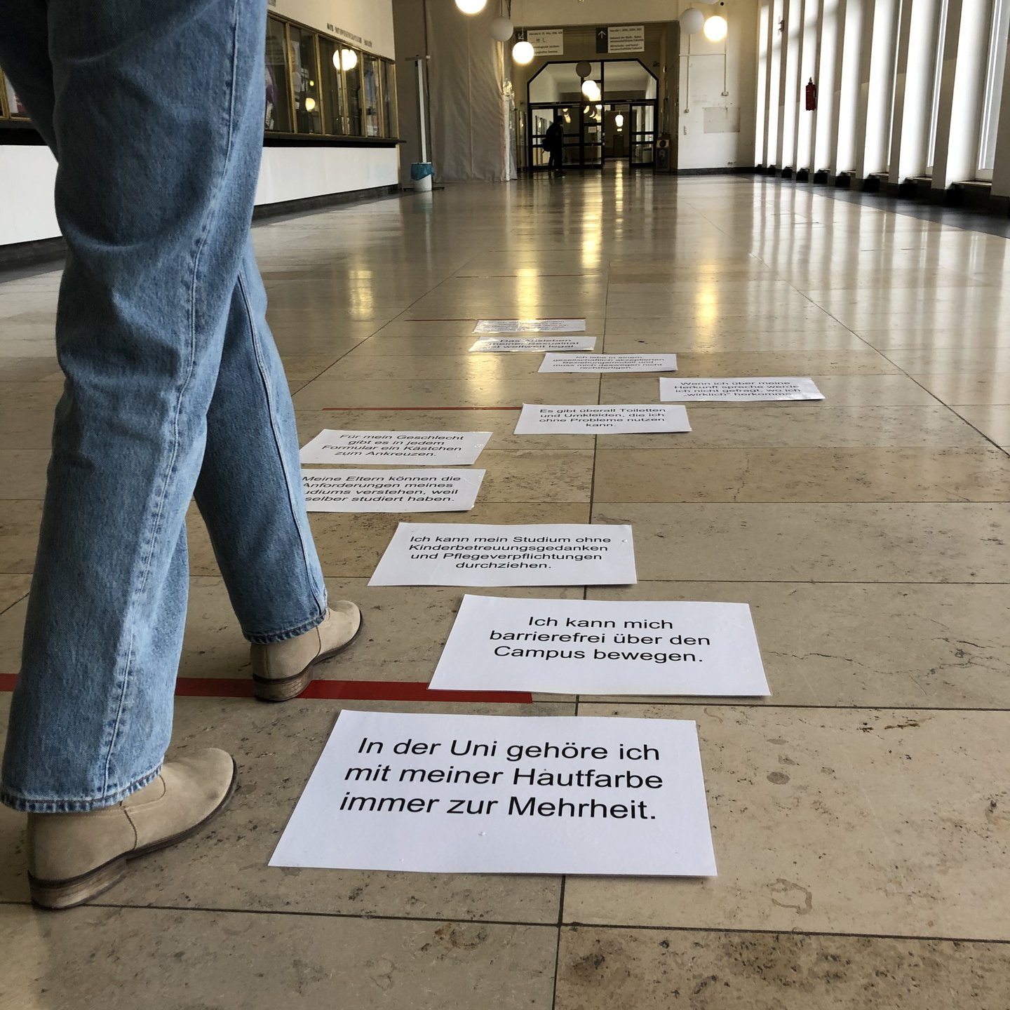 A person walks in the main building of the University of Cologne next to posters that are lying on the floor and form a path. The first three cards each say "at the university I belong to the majority with my skin color", "I can move around the campus without barriers" and "I can complete my studies without having to worry about childcare or care obligations". The other seven cards also contain text that is less legible. A person walks through the building in the background.