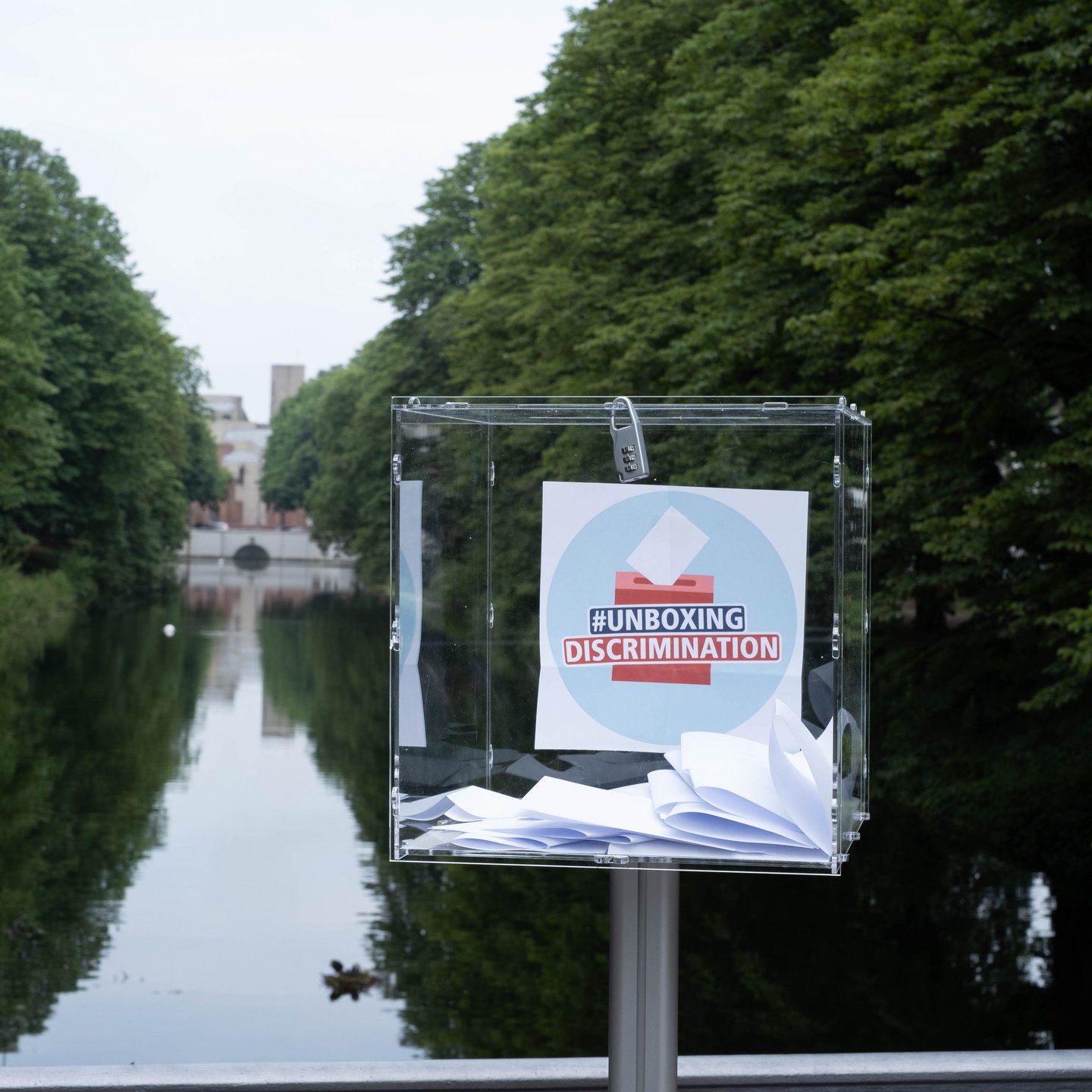 A transparent plexiglass box on a stand stands on the Clarenbachkanal at the Faculty of Human Sciences at Cologne University. Inside the box is the logo of the campaign #unboxingdiscrimination. There are also some white slips of paper collected in the box.