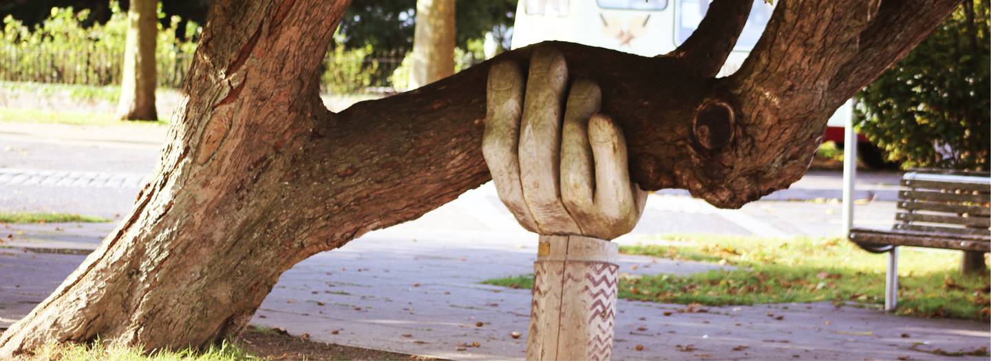 Next to a path, a statue of a large hand is holding up a large tree branch.