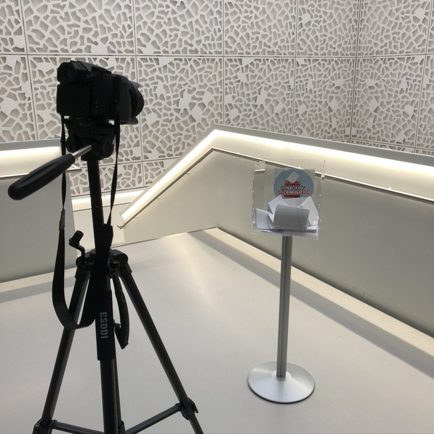 Eine Kamera auf einem Stativ zeigt auf eine Plexiglassbox auf einem silbernen Ständer, die im Treppenhaus des SSC der Uni Köln steht. In dieser Box sind zahlreiche weiße Zettel und das Logo der Kampagne #unboxingdiscrimination.