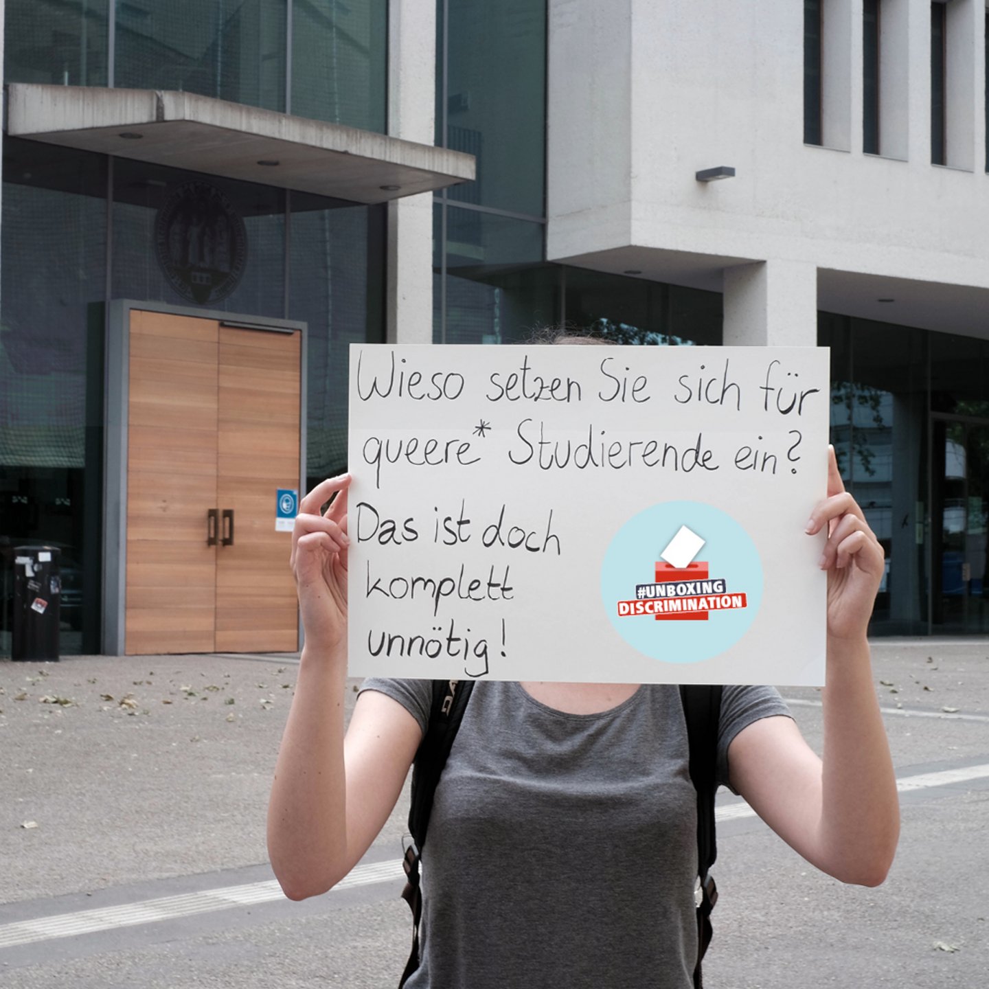 Eine Person steht vor dem Seminargebäude der UzK mit einem Schild vor dem Gesicht mit dem Logo der Kampagne #unboxingdiscrimination und der Aufschrift: Und dafür engagieren Sie sich? Das ist doch komplett unnötig?