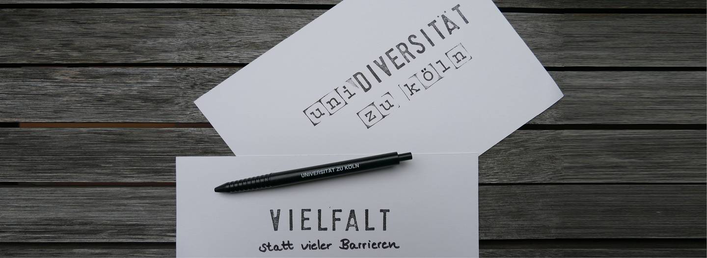Zwei Karten liegen mit einem Stift auf einem Holztisch. Auf der oberen Karte steht 'Uni-Diversität zu Köln', auf der unteren 'Vielfalt statt vieler Barrieren'. 