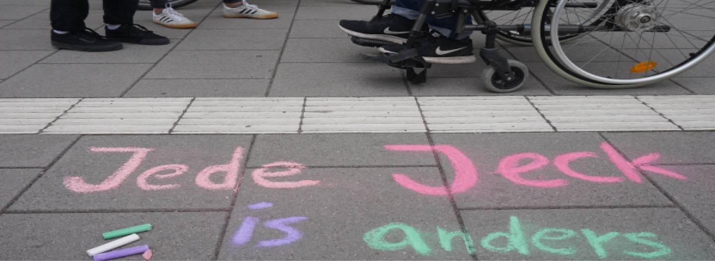 Mit bunter Kreide steht 'Jede Jeck is anders' auf den Albertus-Magnus-Platz geschrieben, auf dem ein Rollstuhlfahrer sitzt.