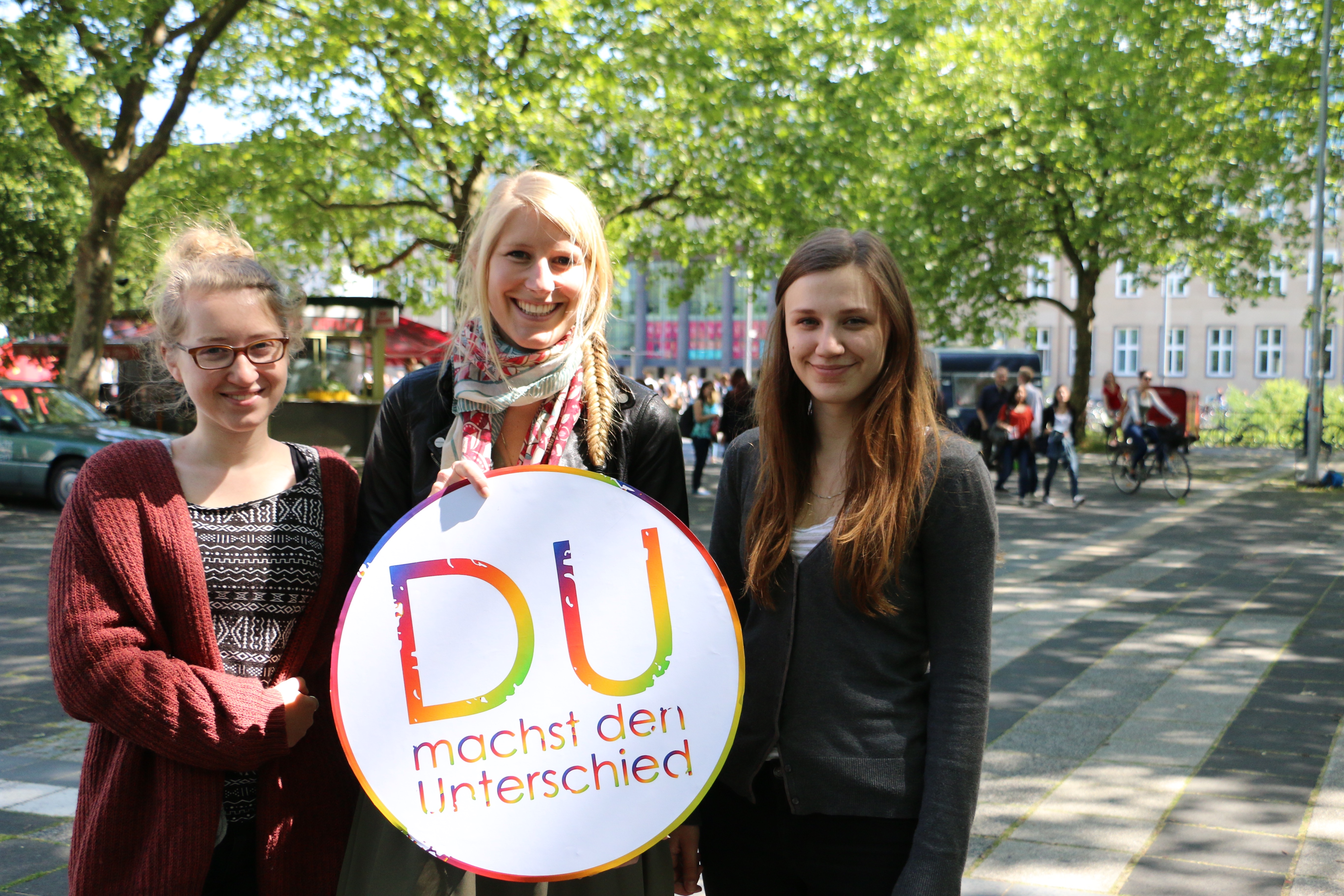 Drei Studentinnen halten gemeinsam auf dem Albertus Magnus Platz das Logo der Themenwoche "Du machst den Unterschied"